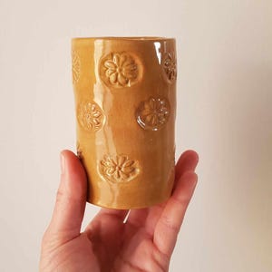 Op de afbeelding: Een cilindervormige keramische vaas in een warme, goudbruine kleur. De vaas heeft reliëf bloemmotieven rondom de buitenkant. De gladde, glanzende afwerking voegt elegantie toe aan dit decoratieve stuk. Ideaal voor bloemen.