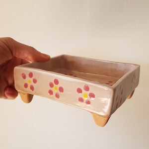 Könnte beinhalten: Eine rechteckige Keramikschale mit hellrosa Glasur, verziert mit rosa und gelben Blumenmustern. Die Schale steht auf vier kleinen, abgerundeten Füßen. Der Innenraum ist in einem dunkleren Rosaton gehalten.