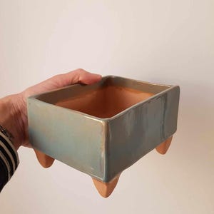 Op de afbeelding: Een vierkante keramische plantenbak met een lichtblauwe glazuur en drie kleine, bruinachtige voetjes. De binnenkant van de plantenbak is warm terracotta van kleur. Ontworpen voor binnen of buiten.