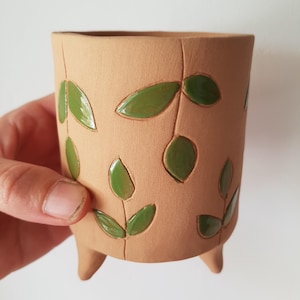 Op de afbeelding: Een cilindervormige terracotta plantenpot met drie korte pootjes, voorzien van een matte afwerking en een uitgesneden patroon van groene geglazuurde bladeren aan stengels. Het oppervlak heeft een natuurlijke aardse textuur met glans.