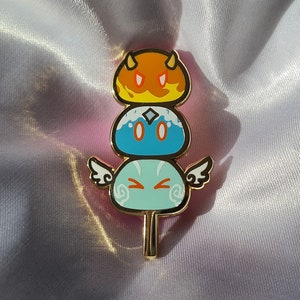 Puede incluir: Un pin de color dorado con tres mochi apilados, cada uno con una cara y un color diferente. El mochi superior es naranja con una cara roja, el mochi del medio es azul con una cara blanca y el mochi inferior es verde con una cara blanca. Los mochi están en un palo.