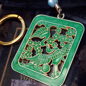 Könnte beinhalten: Ein grüner Metall-Anhänger mit goldener Umrandung, der zwei Kraniche in einem stilisierten Design mit Kirschblüten zeigt. Der Anhänger ist an einem goldenen Ring und einer Perle befestigt.