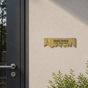 Puede incluir: Un letrero rectangular de metal dorado con acabado cepillado, que presenta un gráfico negro de una torre y la silueta de un horizonte urbano. El texto Studio Tecnico Geom. Marco Rossi aparece en una fuente negra sans-serif clara y legible.