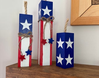 May include: Three wooden blocks painted in red, white, and blue with stars and stripes, resembling fireworks. The blocks are arranged on a wooden shelf.