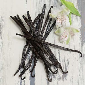 May include: A cluster of dark brown vanilla beans with a few white and pink flowers and green leaves on a distressed, white wooden surface. The vanilla beans are long and slender, with a slightly curved shape.