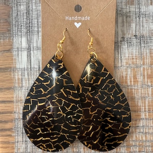 May include: A pair of handmade teardrop-shaped earrings with a black and gold crackle pattern. The earrings are on a brown card with the word "Handmade" written on it.
