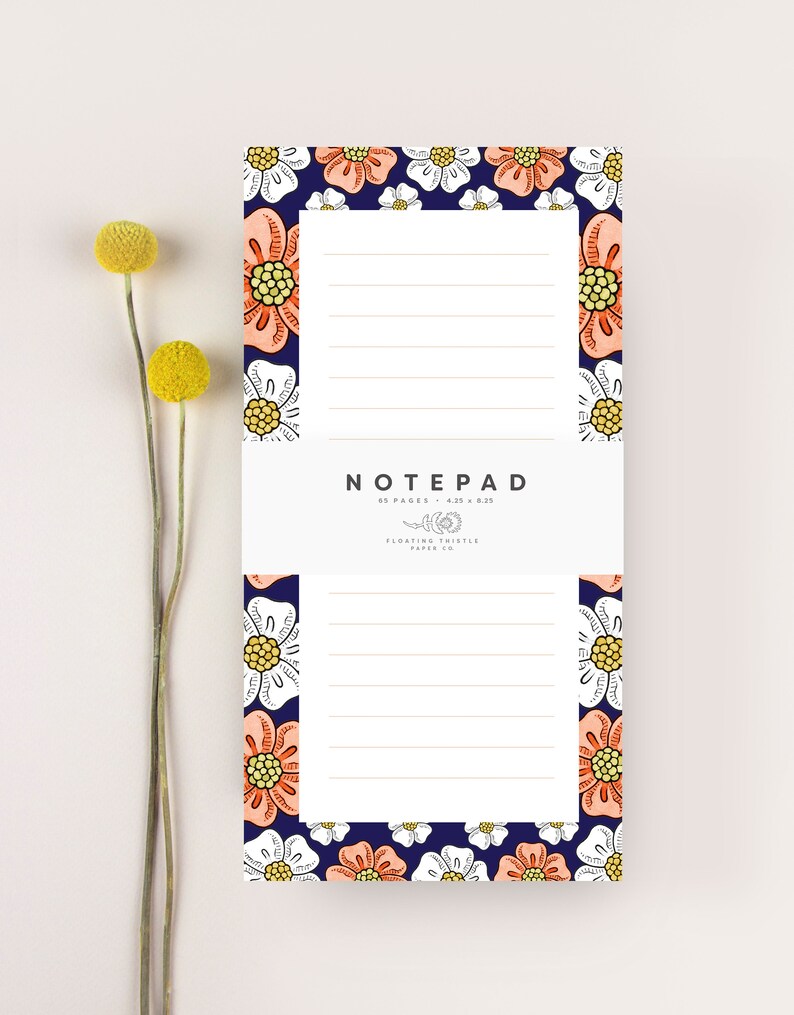 May include: A notepad with a floral design featuring white and peach flowers on a navy blue background. The notepad has 85 pages and measures 4.25 x 8.25 inches. The text "NOTEPAD" is printed in white at the top of the notepad. The text "FLOATING THISTLE PAPER CO." is printed in white at the bottom of the notepad.