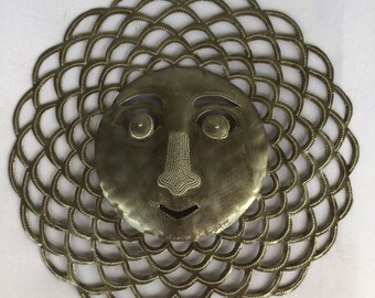 May include: A brass-toned metal sun face wall hanging. The face features large eyes, a beaded nose, and a smiling mouth. The sun's rays are created by a scalloped, openwork design. The wall hanging is mounted on a white wall.