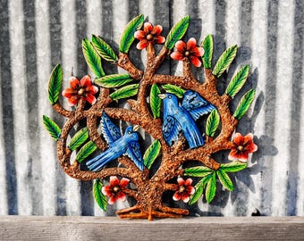 May include: A metal wall art piece depicting a tree with green leaves and red flowers. Two blue birds are perched on the branches. The tree is painted in a rustic style with a gold finish.