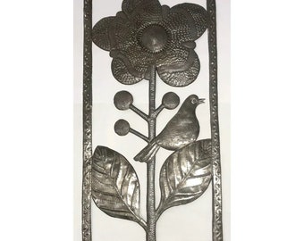 May include: A silver metal wall art piece depicting a stylized flower with a bird perched on a branch. The flower has a large center and three petals. The bird is facing right and has a long tail. The piece is framed with a decorative border.