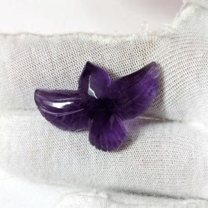 May include: A carved amethyst gemstone in the shape of a stylized flower. The purple gemstone has detailed leaf and petal carvings. The flower is displayed on a white, textured surface.