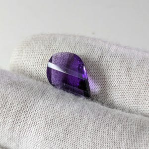May include: A faceted, teardrop-shaped amethyst gemstone. The gem is a deep purple colour with a white, diagonal stripe across the top. The stone is held against a light grey, textured fabric.