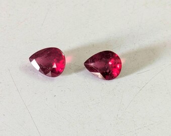 May include: Two faceted, pear-shaped gemstones with a deep red hue. The gems are displayed on a white surface, showcasing their polished facets and internal brilliance. The gemstones are likely rubies, suitable for jewelry.