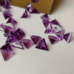 May include: A collection of triangular amethyst gemstones, each with a purple hue and a faceted cut. The gemstones are scattered on a white surface.