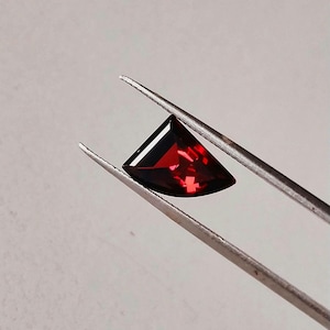 May include: A faceted, deep red gemstone with a triangular, shield-like shape held by a pair of silver-toned metal tweezers. The stone exhibits internal light reflections and a polished surface, showcasing a vibrant, saturated crimson color throughout.