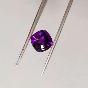 May include: A deep purple cushion-cut gemstone held by tweezers.