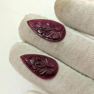 以下が含まれることがあります： 深紅色の涙型の彫刻が施された2つの宝石。各宝石は複雑な花の模様が特徴です。宝石は白く質感のある表面に表示され、その色と詳細を強調しています。