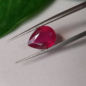 Può includere: Una pietra preziosa di rubino a forma di pera tenuta da una pinzetta. Il rubino è di colore rosso intenso e ha una sfaccettatura.