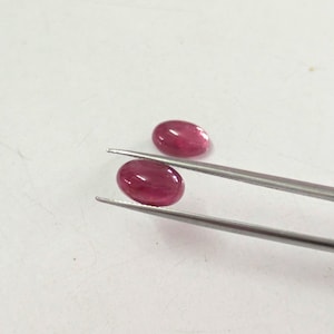 May include: Two oval-shaped, deep pinkish-red gemstones with a smooth, polished cabochon finish. One gemstone is held securely by a pair of silver-toned metal tweezers, while the second gemstone rests nearby on a plain, light-coloured surface.