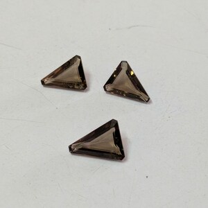 May include: Three triangular, faceted gemstones in a smoky brown color. The gems are cut with sharp angles and appear to be transparent. They are arranged on a light-colored surface.