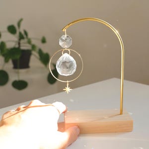 Peut inclure: Un support en métal doré avec une base en bois soutient un prisme en cristal suspendu à un anneau doré. Le prisme est une sphère transparente et facetée avec un plus petit cristal faceté au-dessus. Un charme d'étoile dorée pend sous la sphère.