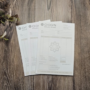 May include: Three white order forms with the text "CHOGAN BESTELLUNG/RECHNUNG" at the top. The forms have a black floral design and are on a wooden surface.