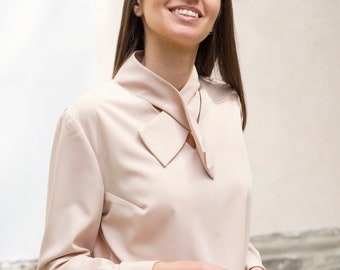 Könnte beinhalten: Eine hellbeige Bluse mit einem einzigartigen Kragendesign und langen Ärmeln. Die Bluse hat eine Schleife am Ausschnitt und wird mit einem passenden Rock kombiniert. Das Outfit ist in einer neutralen Farbe gehalten und für verschiedene Anlässe geeignet.