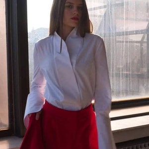 Puede incluir: Una mujer con una camisa blanca de manga larga con un cuello único y pantalones cortos rojos. La camisa tiene puños anchos y un diseño cruzado. Los pantalones cortos son de cintura alta y de color rojo vibrante. El atuendo se combina con medias negras.