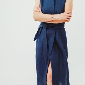 May include: A sleeveless, navy blue dress with a wrap-style waist tie and a high collar. The dress features a thigh-high slit and is made of a textured fabric. The model is wearing a silver bracelet.