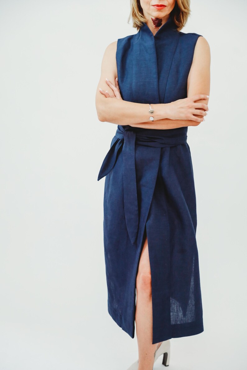May include: A sleeveless, navy blue dress with a wrap-style waist tie and a high collar. The dress features a thigh-high slit and is made of a textured fabric. The model is wearing a silver bracelet.