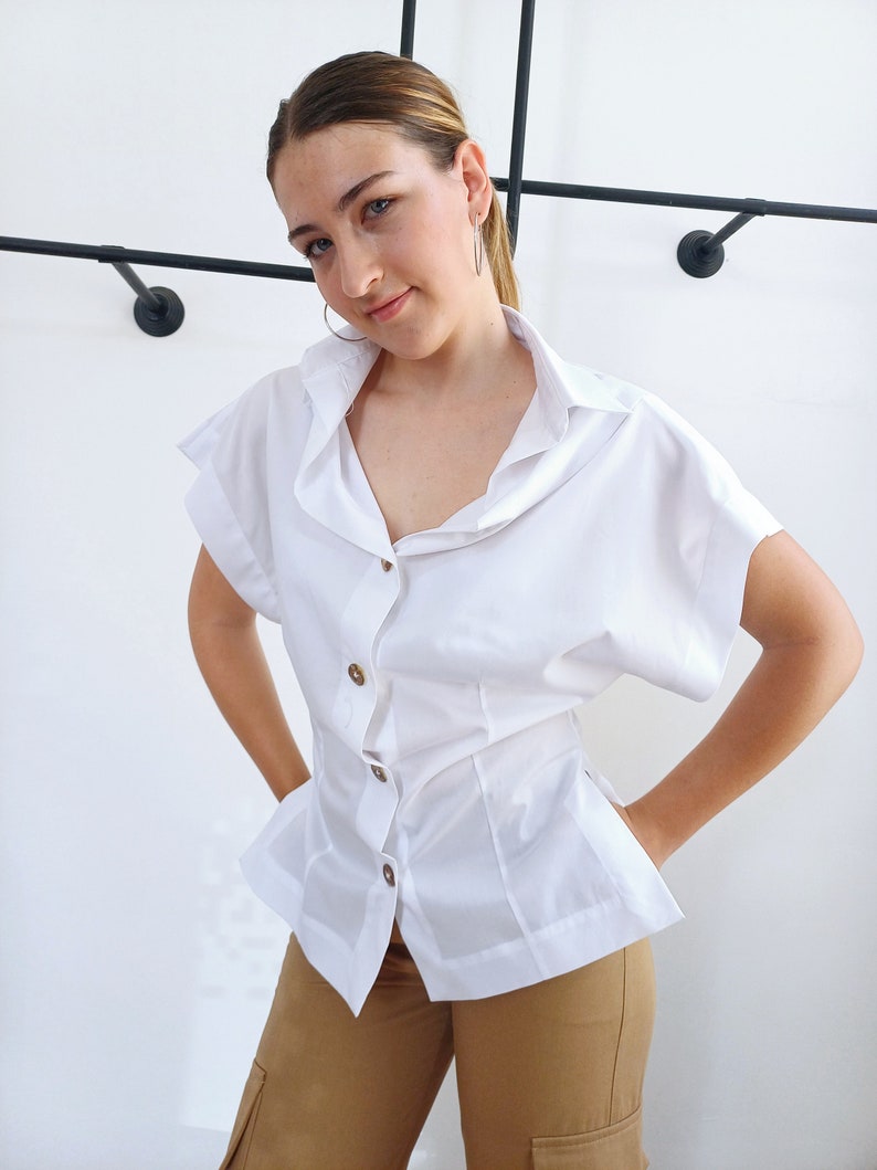 May include: A white short-sleeved blouse with a button-down front and a unique collar design. The blouse has a fitted waist and is paired with tan cargo pants. The outfit is simple and stylish.