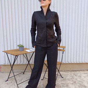 May include: A black linen suit consisting of a long-sleeved shirt with a high collar and wide-leg pants. The outfit is worn with black sandals. A small wooden table and chair are in the background.