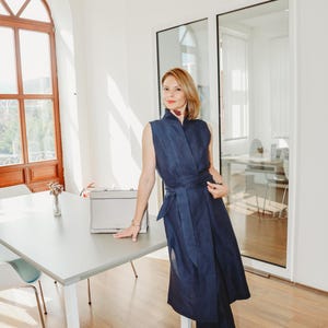 May include: A woman wearing a navy blue sleeveless dress with a matching belt and pants. The dress has a high collar and a wrap-around design. She is standing next to a gray table with a silver briefcase on it.