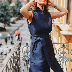 May include: A sleeveless navy blue dress with a matching belt is worn by a person on a balcony. The dress has a high neckline and a flowing skirt. The person is wearing sunglasses and has their arms raised. The background shows a city scene.