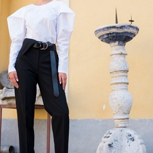 Puede incluir: Una mujer viste una blusa blanca de manga larga con mangas estructuradas y pantalones negros de cintura alta. Un cintur&oacute;n negro con hebilla plateada ci&ntilde;e la cintura. Lleva tacones negros. Un pilar de piedra decorativo en el fondo. Look elegante.