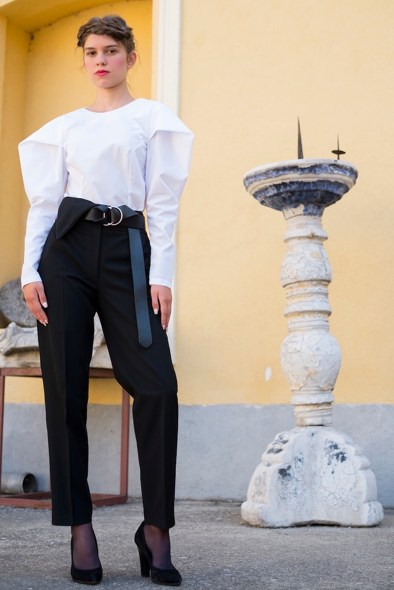 Puede incluir: Una mujer viste una blusa blanca de manga larga con mangas estructuradas y pantalones negros de cintura alta. Un cintur&oacute;n negro con hebilla plateada ci&ntilde;e la cintura. Lleva tacones negros. Un pilar de piedra decorativo en el fondo. Look elegante.