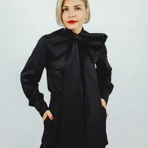 Peut inclure: Une personne porte un chemisier noir en satin avec un grand nœud papillon et des manches longues. Elle porte également un pantalon noir. Le fond est blanc uni.