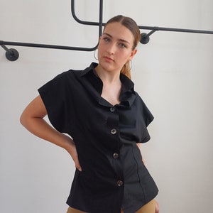 May include: Black button-down shirt with short sleeves. The shirt has a classic collar and is buttoned down the front with large, round buttons. The shirt is paired with tan pants.