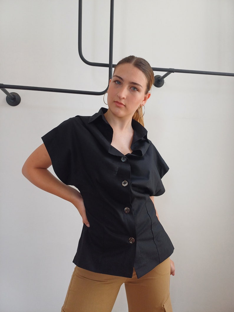 May include: Black button-down shirt with short sleeves. The shirt has a classic collar and is buttoned down the front with large, round buttons. The shirt is paired with tan pants.