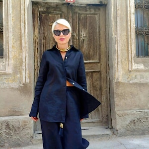 May include: A woman wearing a navy blue linen shirt and matching wide-leg pants. The shirt is unbuttoned at the waist, and a gold necklace is visible. The background features a weathered wooden door with the number 92.
