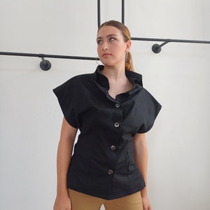 May include: A black button-down shirt with short sleeves. The shirt has a high collar and large buttons down the front. The model is wearing tan cargo pants. The background is a white wall with black metal rods.