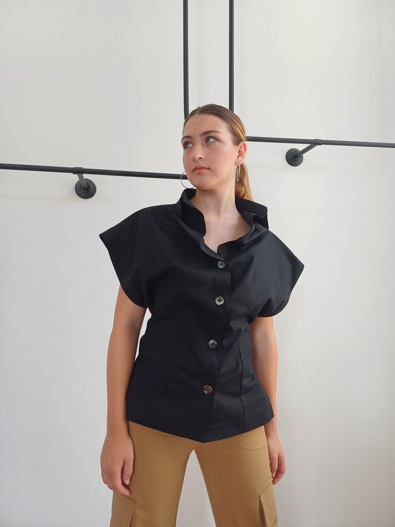 May include: A black button-down shirt with short sleeves. The shirt has a high collar and large buttons down the front. The model is wearing tan cargo pants. The background is a white wall with black metal rods.