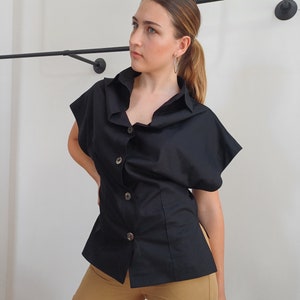May include: A black short-sleeved button-down shirt with a unique collar design. The shirt has a fitted waist and is paired with tan cargo pants. The shirt features large, decorative buttons. The background is a plain white wall.