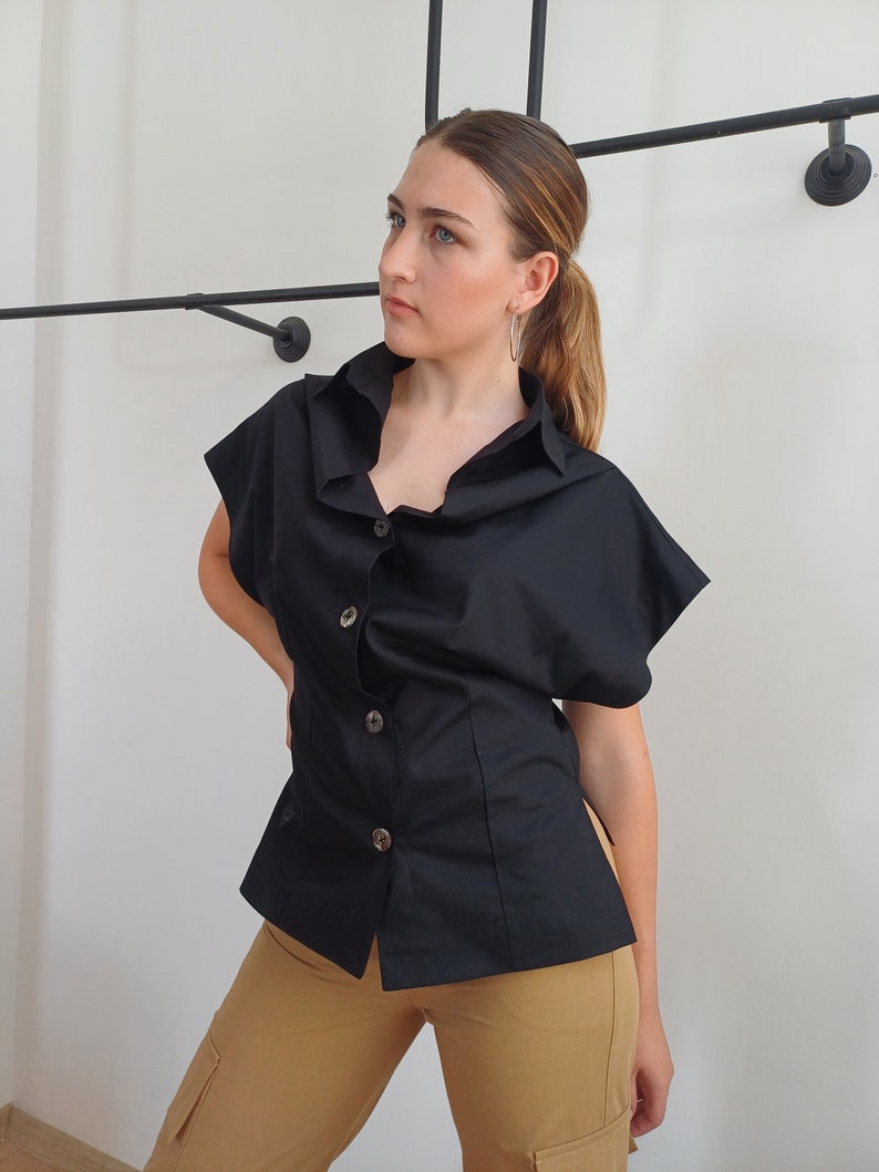 May include: A black short-sleeved button-down shirt with a unique collar design. The shirt has a fitted waist and is paired with tan cargo pants. The shirt features large, decorative buttons. The background is a plain white wall.
