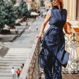 May include: A sleeveless navy blue jumpsuit with a matching belt. The jumpsuit has a high collar and wide-leg pants. The person is wearing light-colored shoes and sunglasses. The background shows a city street with buildings and people.