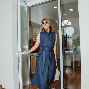 May include: A woman wearing a sleeveless navy blue jumpsuit with a matching belt and sunglasses. She is holding a white handbag and opening a glass door. The outfit has a high collar and a wrap-style design.