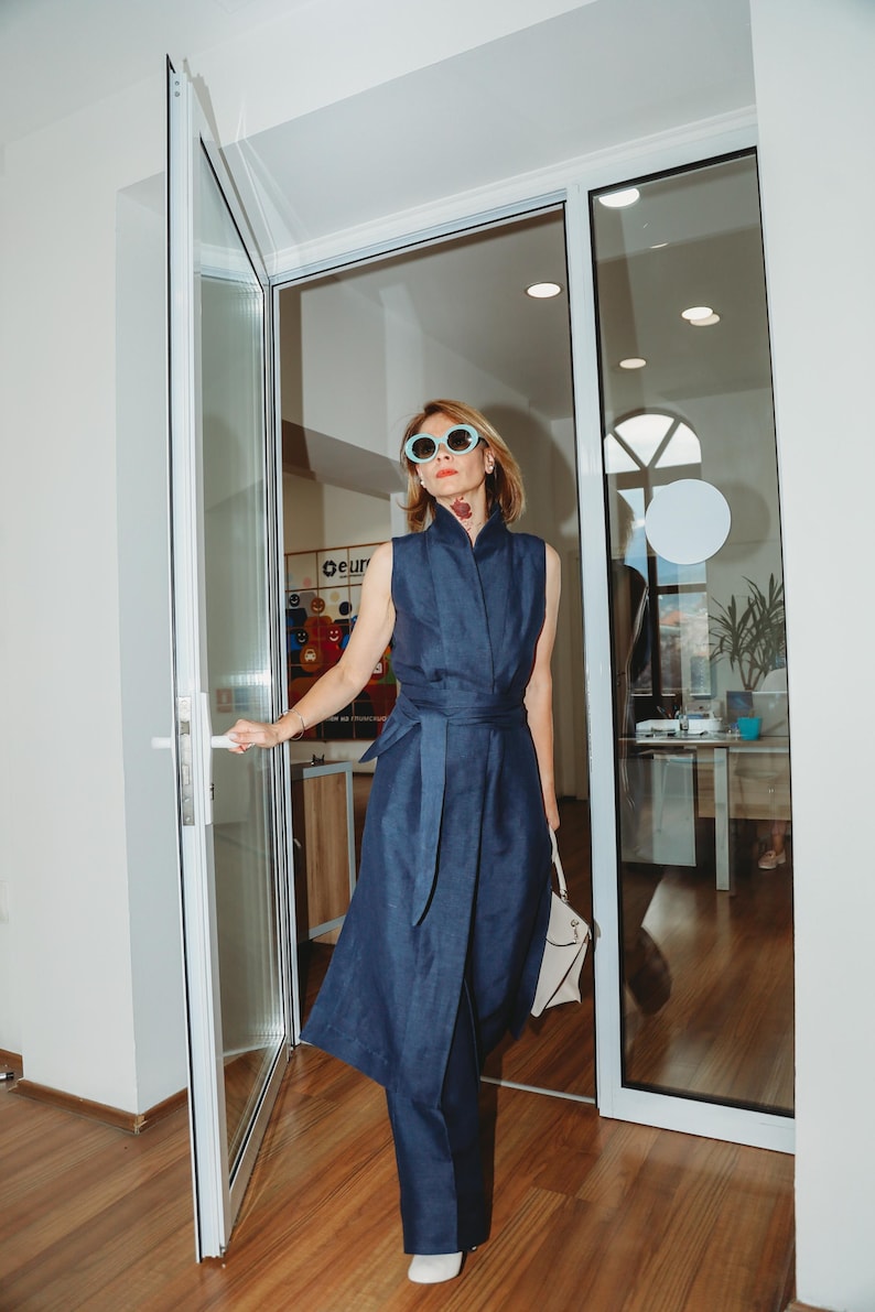 May include: A woman wearing a sleeveless navy blue jumpsuit with a matching belt and sunglasses. She is holding a white handbag and opening a glass door. The outfit has a high collar and a wrap-style design.