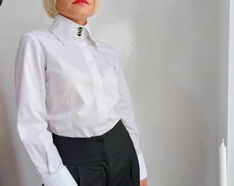 May include: A crisp white long-sleeved button-down shirt featuring a high collar with two prominent black and gold circular buttons, paired with black high-waisted trousers. The shirt has wide cuffs, each secured with a single matching black and gold button.