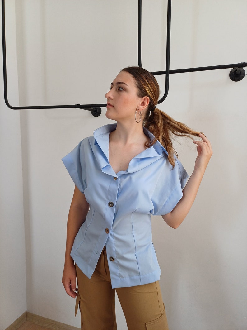 May include: A light blue button-down shirt with short sleeves and a tailored waist. The shirt features a classic collar and is paired with tan cargo pants. The outfit is set against a white wall with a black metal clothing rack.