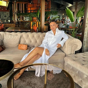 Può includere: Una donna con un abito camicia bianco con spacco alto, seduta su un divano beige trapuntato. Indossa orecchini a cerchio dorati e sandali marroni. Una piccola borsa marrone è sul divano. Due tavoli rotondi con piano in vetro e cornici dorate sono davanti.